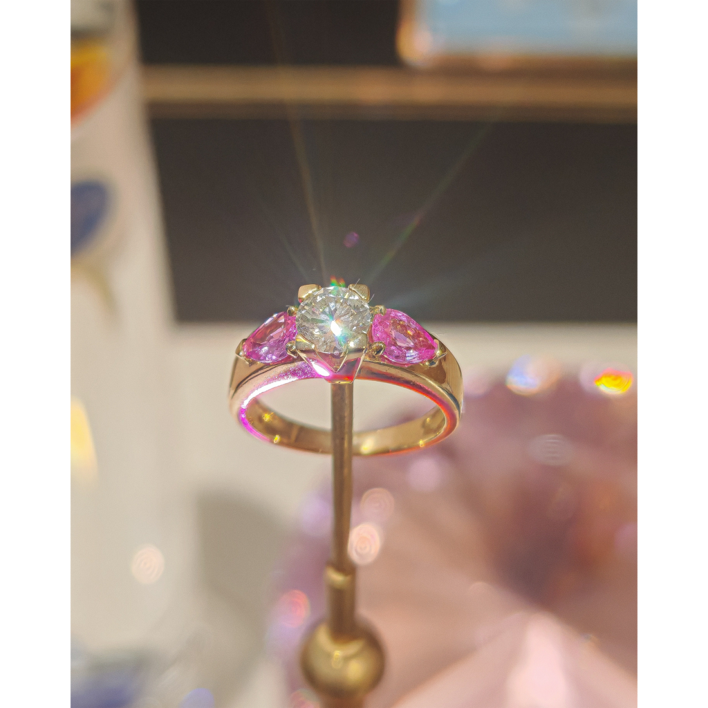 Bague en or jaune 18 carats sertie de deux saphirs roses et d'un diamant central, la lumière du diamant brille et fait une sorte de halo, la bague est posée sur un élément de décor